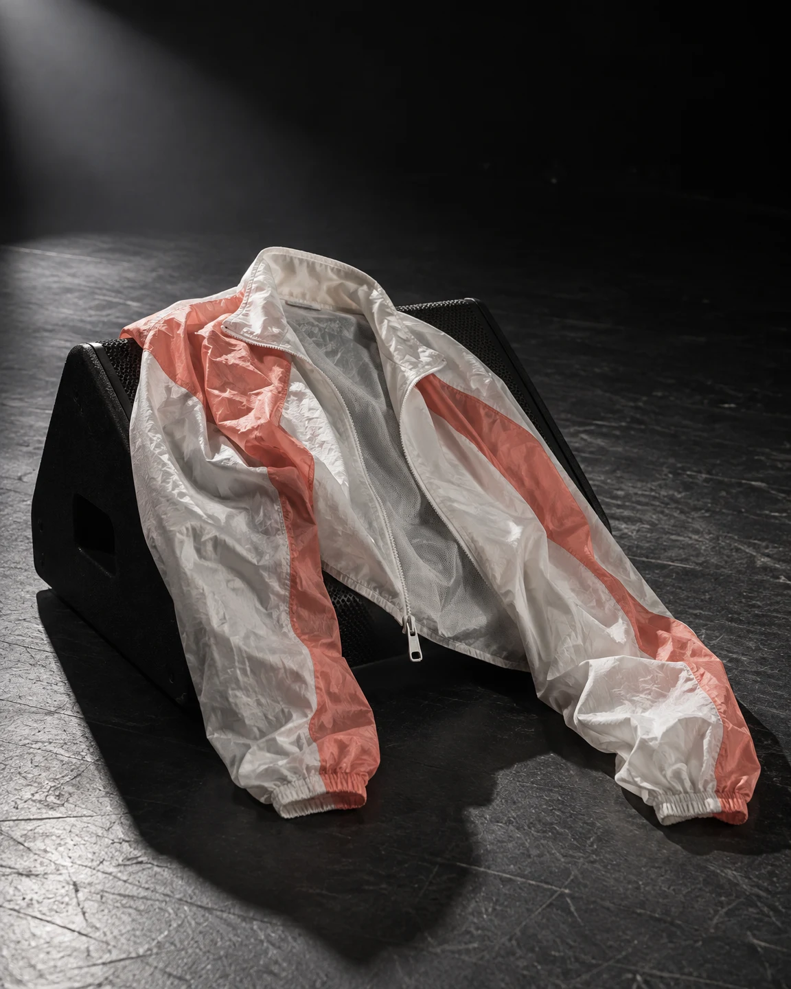 A pale nylon track jacket with coral panels is draped over a black wedge-shaped stage monitor, its open front and sleeves falling toward a scuffed charcoal…