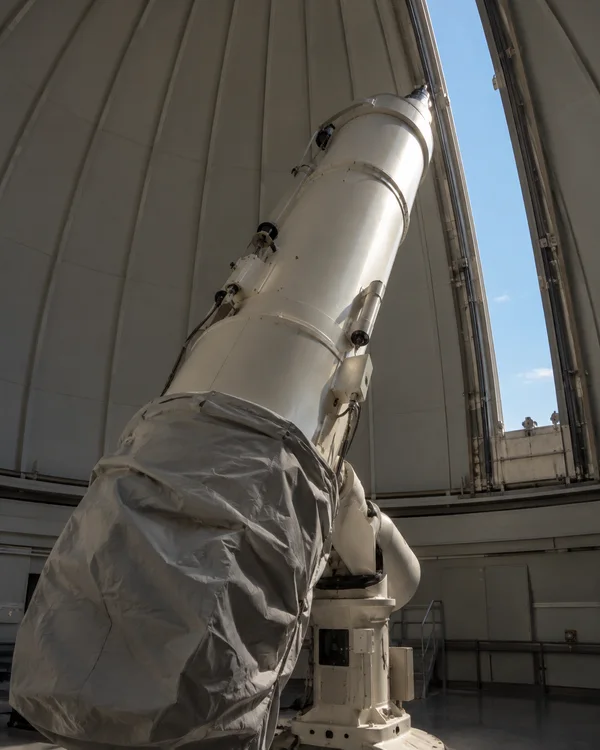 A large off-white telescope rises inside a curved observatory dome, its lower tube partly wrapped in loosely bunched silver-grey fabric.