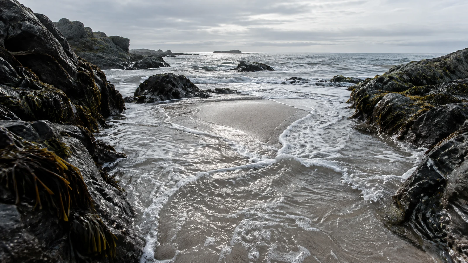 Foamy incoming water winds around a pale sandbar between dark, seaweed-streaked rock ledges on a rugged coast, with a grey sea and cloudy sky on the horizon.