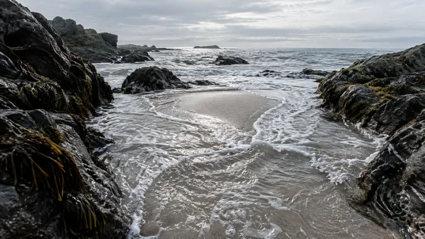 Foamy incoming water winds around a pale sandbar between dark, seaweed-streaked rock ledges on a rugged coast, with a grey sea and cloudy sky on the horizon.