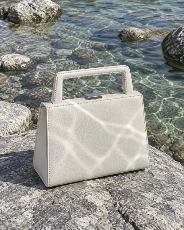 A chalk-white structured leather handbag with a rectangular handle and silver clasp sits on a sunlit grey rock beside clear, shallow water and rounded stones.