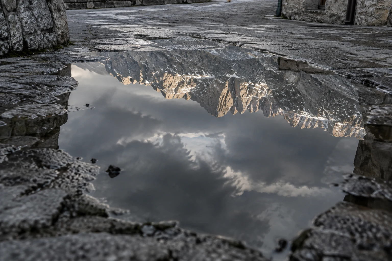 A jagged, warmly lit mountain ridge and dark clouds reflect in a large puddle on a wet stone street, bordered by irregular paving and a stone building wall.