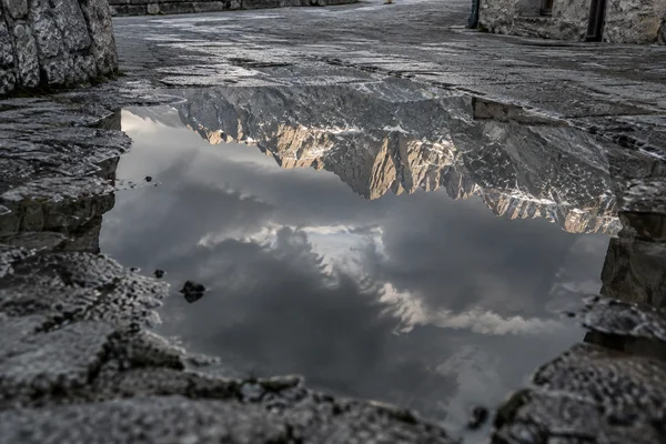 A jagged, warmly lit mountain ridge and dark clouds reflect in a large puddle on a wet stone street, bordered by irregular paving and a stone building wall.