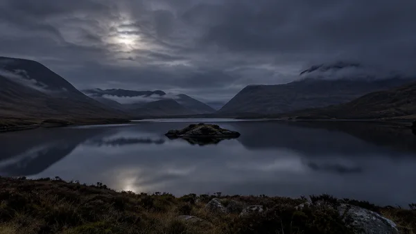 A small rocky islet sits near the center of a still upland lake, reflecting beneath clouded midnight skies in a broad mountain valley; dark heather and rocks…