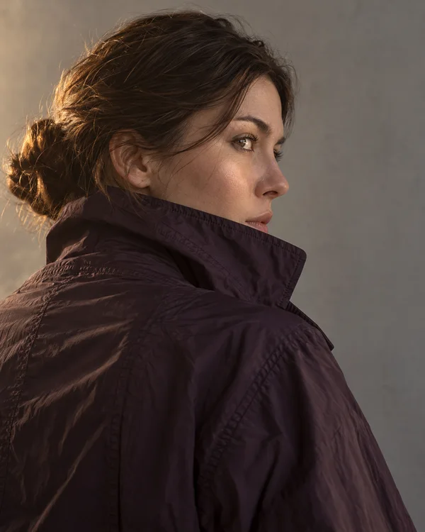 A woman in a dark aubergine high-collar jacket turns to look right in a close rear three-quarter portrait, with warm light catching her face and hair against a…