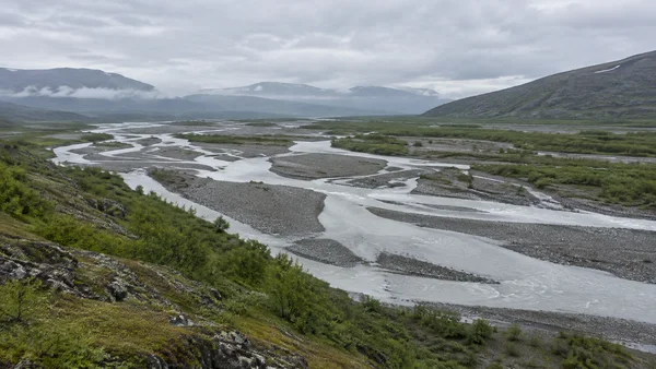 A pale braided river winds across a broad gravel floodplain, bordered by green scrub beneath misty hills and a cloudy sky.