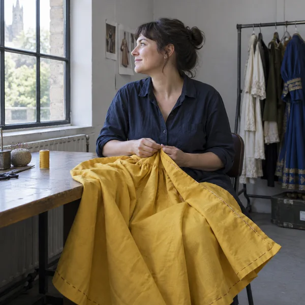 An adult costume maker in a dark blue work shirt sits at a repair table, turned toward a tall window while holding a large yellow skirt with visible basting…