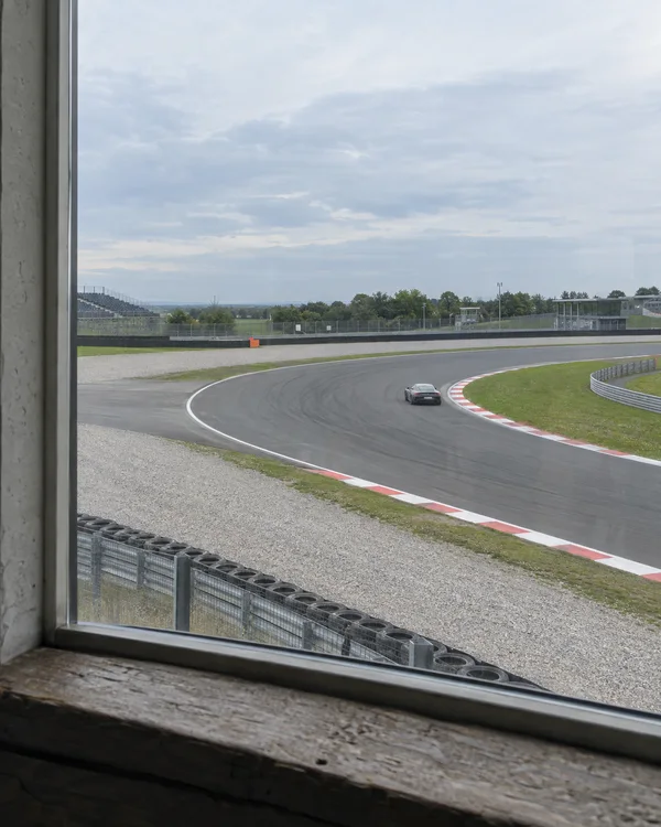 A small grey coupe rounds a broad asphalt bend with red-and-white kerbs at a motor-racing circuit, seen through a large window with a concrete reveal on the…
