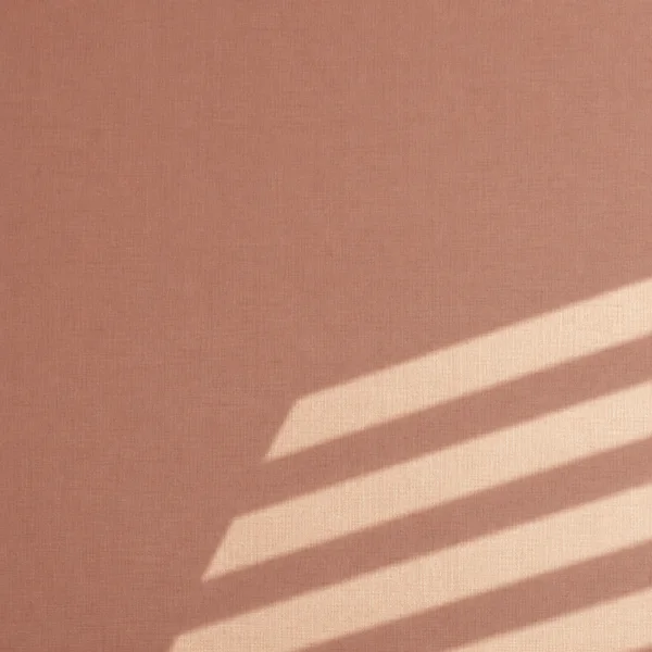A square clay-pink woven textile surface with four broad diagonal ivory light bands entering from the right and occupying the lower-right portion of the frame…