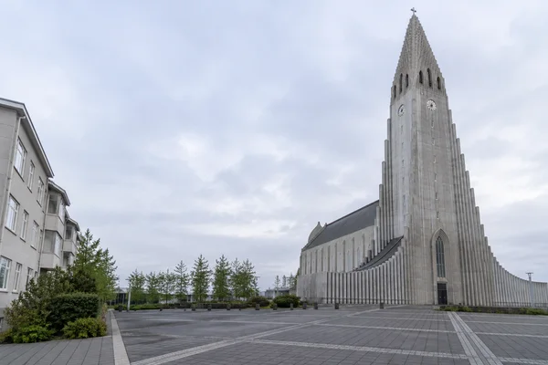 A stepped concrete church tower with clock faces rises beside its adjoining church volume, overlooking a broad paved forecourt under an overcast sky; an…