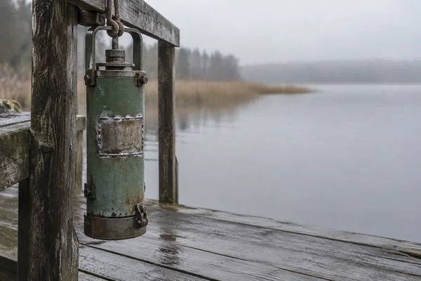 A weathered green cylindrical steel sampler hangs from a rope beside the rail of a damp wooden jetty, its rough rectangular welded patch prominent against a…