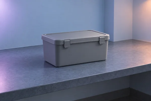 A matte gray rectangular storage box with a fitted lid and two front clip latches sits centered on a slate-blue bench in a bare room with pale blue walls.