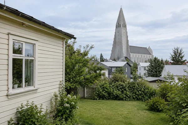 A cream timber cottage with a divided-pane window borders a small garden lawn, with neighbouring rooftops and Hallgrímskirkja rising in the background beneath…