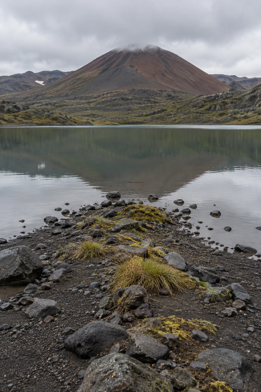 A narrow black volcanic shoreline with scattered basalt rocks and tufts of grass leads into a muted green lake, reflecting a rust-brown cinder cone beneath low…