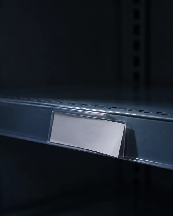 A blank white paper label sits at an angle in a clear plastic holder on a blue-grey metal shelf, lit by cool light against a dark, softly blurred interior.