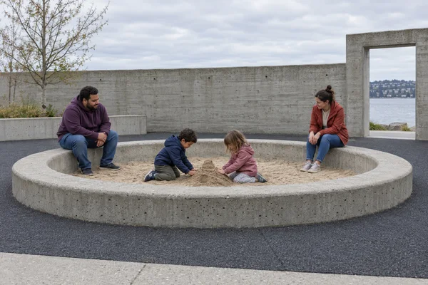 Two children kneel in a circular concrete-rimmed sandpit, shaping a mound while two adults sit on the rim.