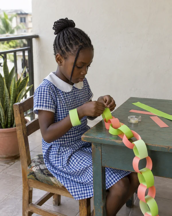 A school-age child in a blue-and-white checked dress sits at a painted wooden table, joining lime-green and coral paper loops; a broad paper loop circles one…