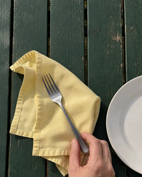 Overhead view of a pale-yellow folded napkin and a stainless-steel fork on a worn dark-green slatted table, beside part of a white plate.