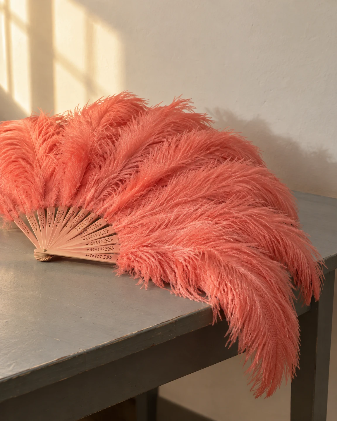 A coral-pink feather fan lies partly open on a grey wooden table, its long plumes spilling over the front edge beneath a sunlit cream wall.