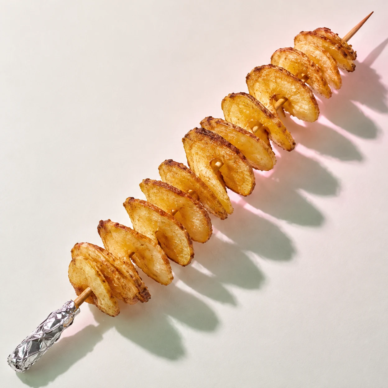 A golden spiral-cut fried potato on a wooden skewer runs diagonally across an off-white tabletop, with a crumpled foil grip at the lower end and long…