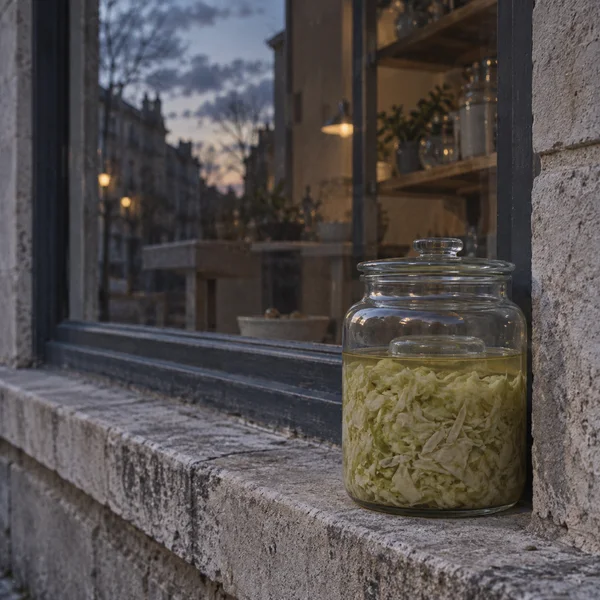 A clear lidded glass jar packed with shredded pale cabbage and brine rests on a weathered stone window ledge.