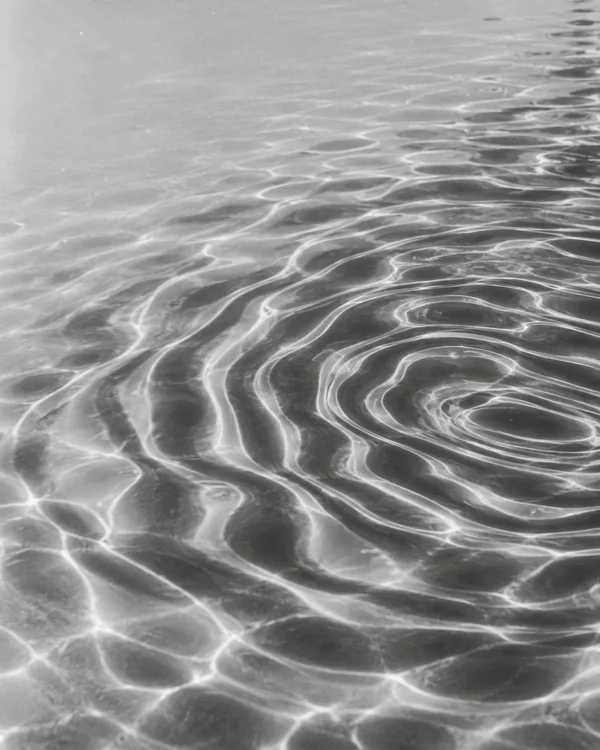 Black-and-white close view of rippling water, with nested oval reflections and sinuous bands across the lower frame and a calmer pale area at upper left.