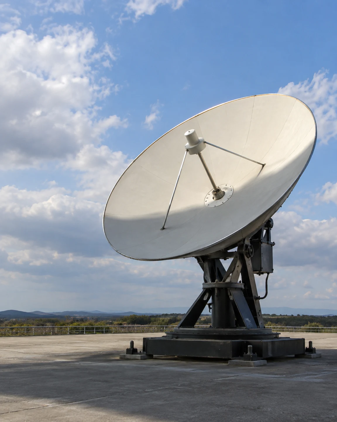 A large ivory parabolic antenna dish on a dark metal pedestal stands on a concrete pad beneath a partly cloudy blue sky, with a low wooded horizon in the…