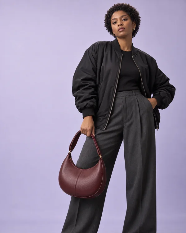 A woman with short natural coils poses against a pale lilac studio backdrop in a black bomber and charcoal wide-leg trousers, holding a textured oxblood…