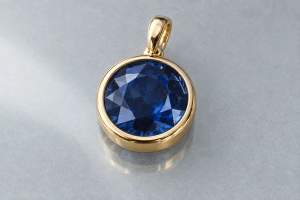 A round deep-blue faceted gemstone pendant in a polished yellow-gold bezel with an integrated oval bail rests on a pale grey surface, with a faint reflection…