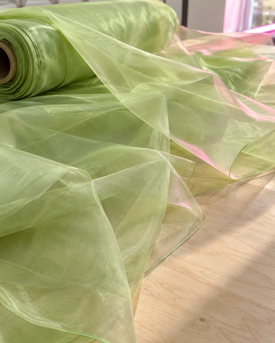 A roll of pale leaf-green sheer fabric spills into translucent folds across a pale plywood table, with pink light catching several edges.
