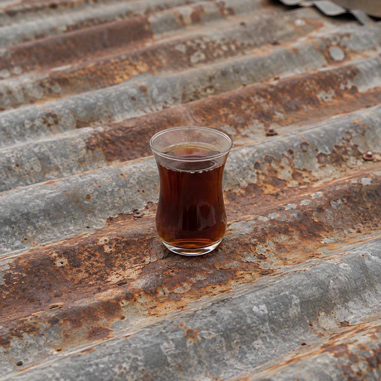 A small clear glass of dark tea with faint wisps of steam sits on a weathered corrugated metal sheet mottled with orange-brown rust and grey galvanised ridges.