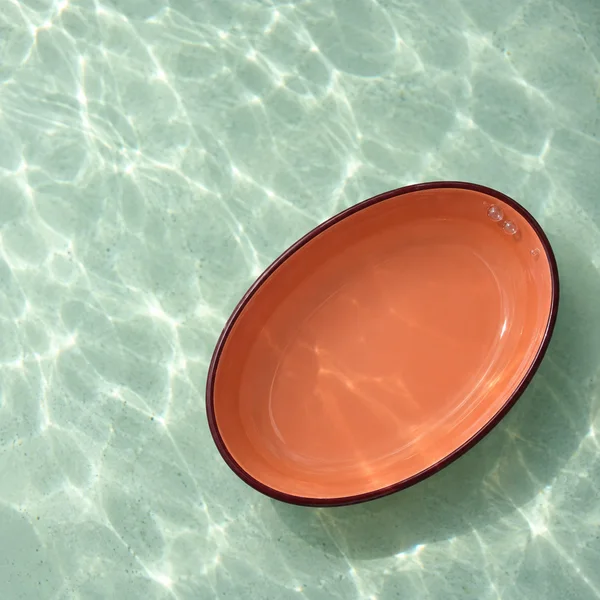 An oval coral-orange enamel bowl with a narrow dark red rim rests diagonally on a pale mint floor beneath clear water.