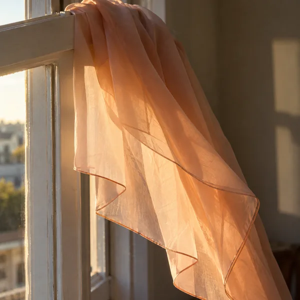 A translucent peach scarf drapes diagonally over a pale window frame, glowing in warm sunlight against a shadowed interior.