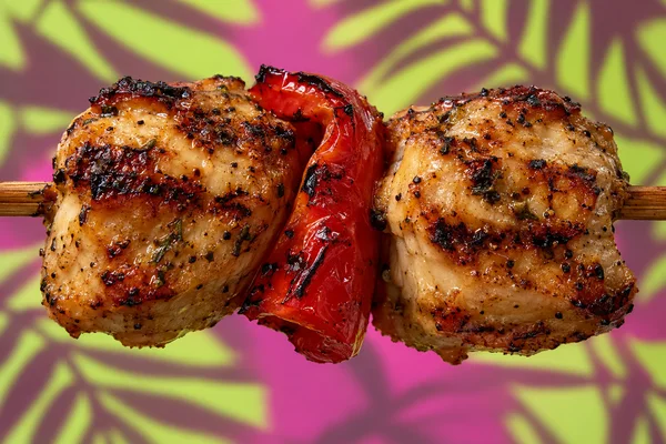 Two large grilled chicken pieces with dark char marks sit on either side of a folded red pepper strip, threaded on a horizontal bamboo skewer against a magenta…