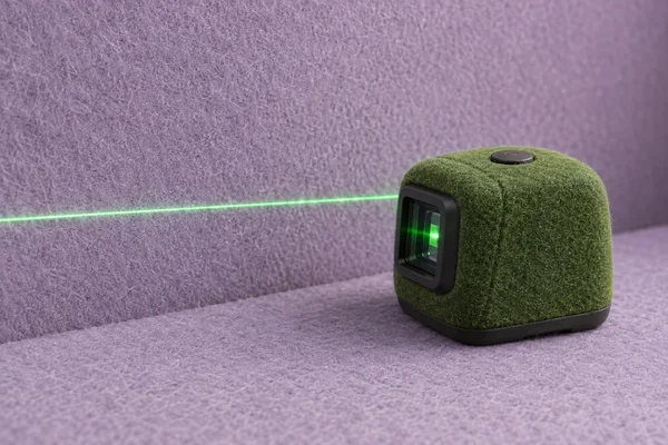 A compact moss-green felt-covered laser level sits on a lilac felt surface against a matching wall, with its dark optical window and top button visible.