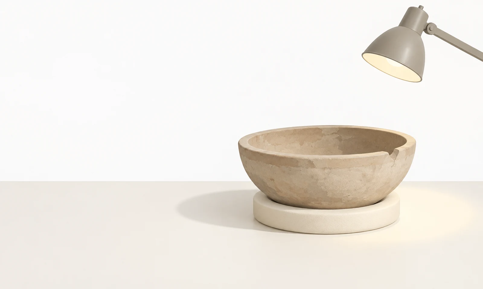 A shallow pale stone pottery bowl with a chipped rim rests on a cream support ring on an ivory tabletop, lit by a muted grey articulated lamp entering from the…