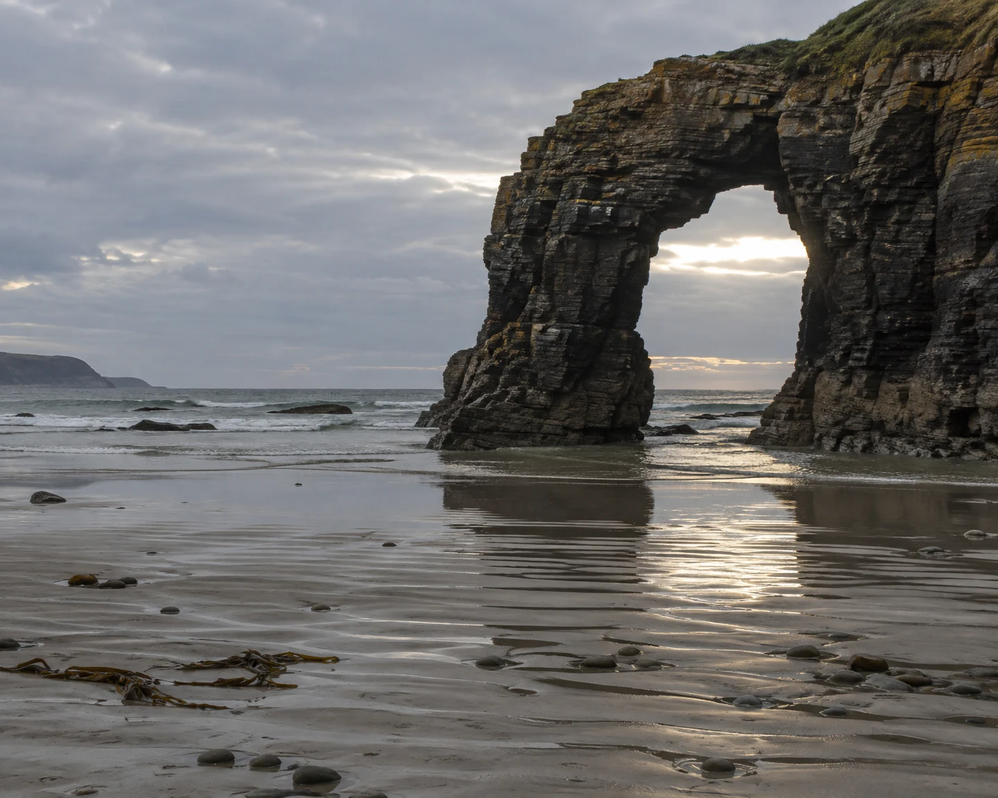A dark, layered sandstone sea arch rises from a wet beach, framing a bright break in the cloudy sky; its light reflects across rippled sand scattered with…