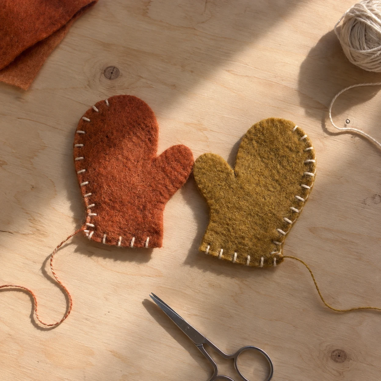 Overhead view of rusty orange and muted ochre felt mitten ornaments on a pale wooden table, with cream edge stitching, loose threads, steel scissors, a ball of…