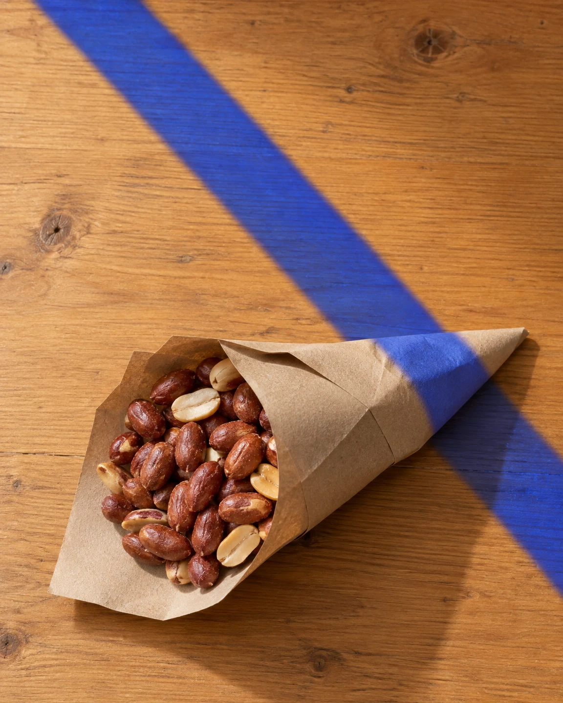 An open brown paper cone filled with reddish skin-on peanuts and pale split nuts lies diagonally on a honey-coloured wooden tabletop.