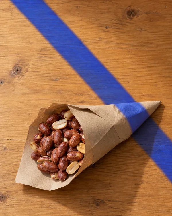 An open brown paper cone filled with reddish skin-on peanuts and pale split nuts lies diagonally on a honey-coloured wooden tabletop.