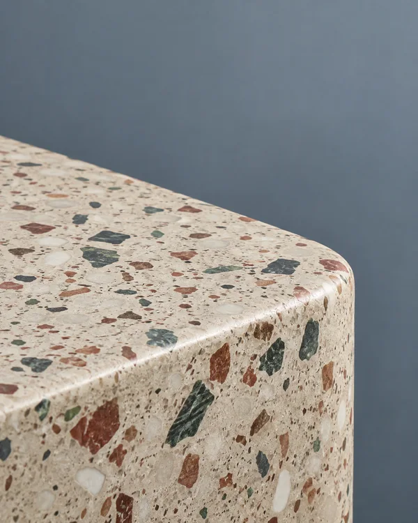 Close-up of a warm off-white terrazzo counter with rust, green and white aggregate, showing its thick fascia and generously rounded corner against a blue-grey…