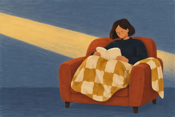 A woman with a dark bob sits curled in a rust-coloured armchair, holding an open blank book beneath a mustard-and-cream checked blanket.