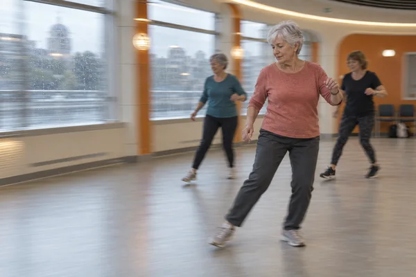 An older woman with short silver hair, a muted coral top, charcoal trousers and sneakers takes a lateral step in a bright hall.