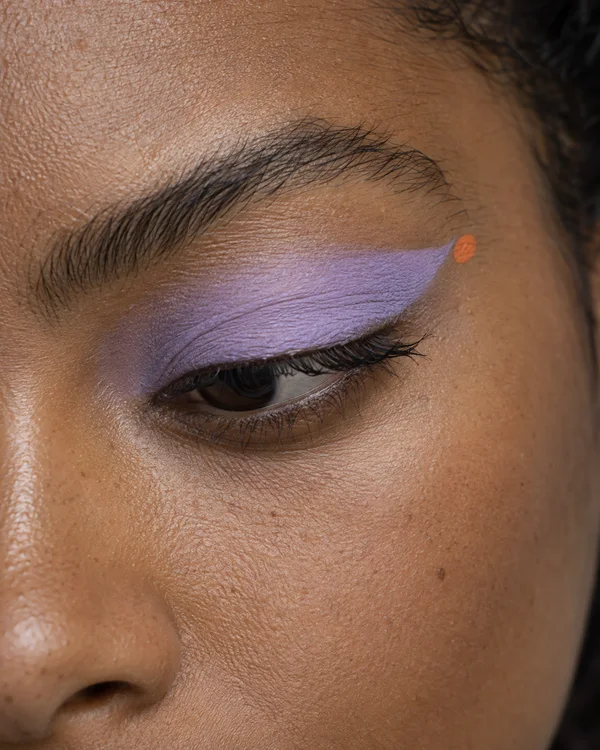 An extreme close-up of a brown-skinned model looking downward, with a broad lilac eyeshadow sweep and a small orange dot beside its outer tip; the eyebrow…