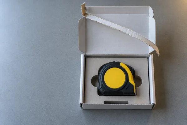 Overhead still life of a yellow-and-black tape measure seated in a fitted insert inside an open white corrugated mailer on a grey tabletop.