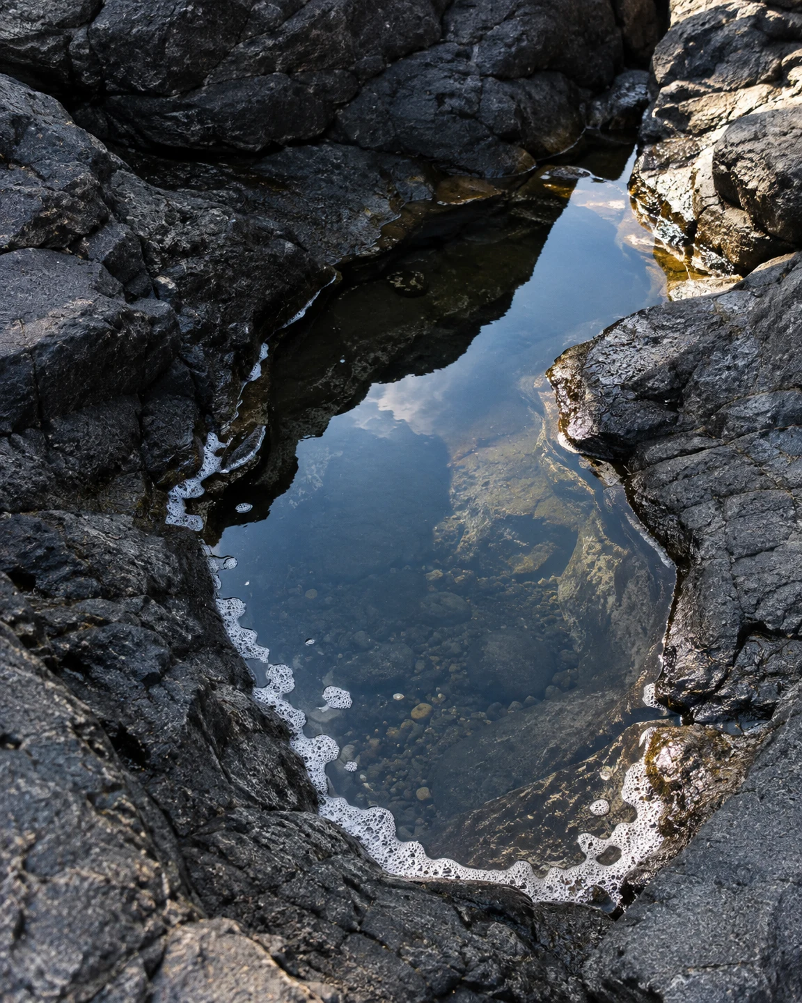 An elongated tidal pool bordered by dark, irregular coastal rocks reflects blue sky and clouds, with submerged pebbles visible through the water and sea foam…