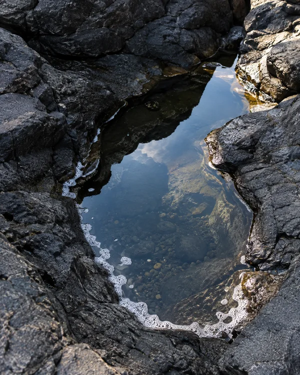 An elongated tidal pool bordered by dark, irregular coastal rocks reflects blue sky and clouds, with submerged pebbles visible through the water and sea foam…