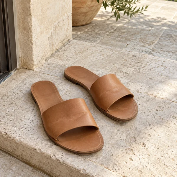 Two matching tan leather slide sandals rest on a pale stone threshold beside a doorway, with a planter and leaves visible on the sunlit terrace beyond.