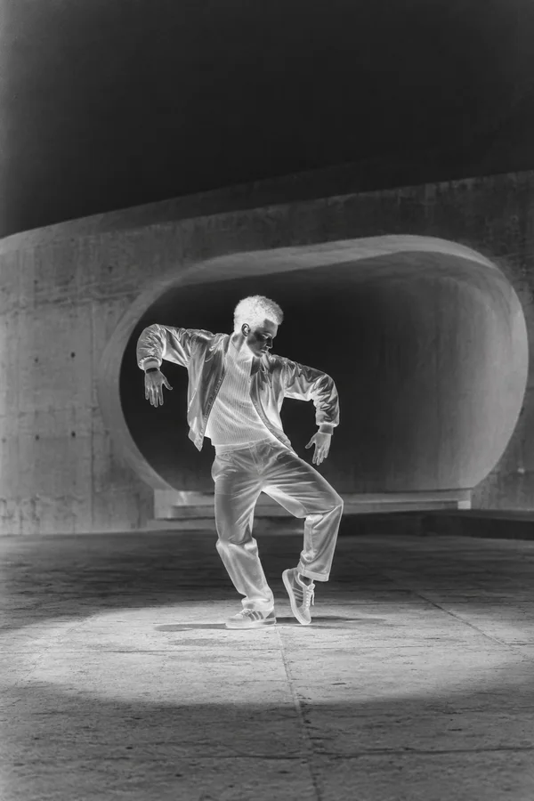 A full-body dancer in a bright jacket, ribbed sweater, loose trousers and sneakers performs a bent-knee pose on a concrete plaza.