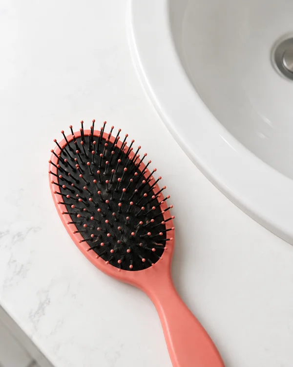 A coral-pink paddle hairbrush lies bristle-side up diagonally on a white counter beside a curved white basin with a metal drain.
